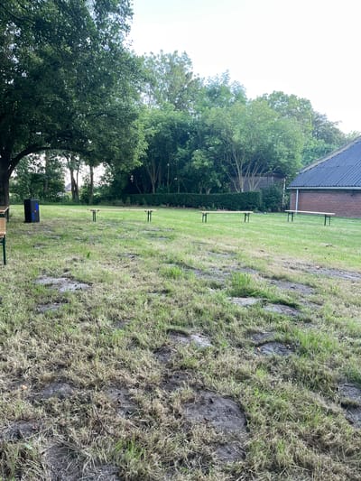 This is an outdoor, eye-level, medium shot of a grassy field in Uithuizen, Netherlands. The image is oriented vertically, with the sky at the top left and the ground occupying the right side of the frame. The field is unevenly mowed, with patches of long, dry grass mixed with darker, muddy areas. Several black metal benches with wooden seats are spaced evenly across the mowed section of the field. In the background, a building with a dark roof and brick facade is partially visible, with trees and a hedge lining the property. The sky is overcast and bright, suggesting a daytime shot under cloudy weather conditions. No people are visible in the frame.