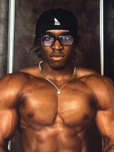 A close-up, waist-up portrait of a muscular Black man wearing glasses and a black beanie. He has a silver chain with a clover-shaped pendant. The background appears to be a textured, dark wall. The lighting is dramatic, highlighting his physique. The man is positioned centrally in the frame, facing the camera directly. There is no identifiable indication of location in Ifite Awka, Nigeria, or any other specific geographical context.