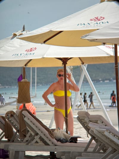 The image depicts a beach scene at Pax Ana Doc Let Resort in Ninh Hoa City, Vietnam. In the foreground, several empty lounge chairs with towels draped over them are arranged on the sand. Behind the chairs, a woman in a yellow bikini stands under a large beach umbrella, wearing sunglasses and a straw hat. She is posing with her hand on her hip. To the left of the woman, a woven basket hangs from the umbrella's pole. In the background, the turquoise ocean stretches out, with several boats visible on the water and a hazy shoreline with buildings. People are scattered across the beach and in the water. The sky is a light blue.
