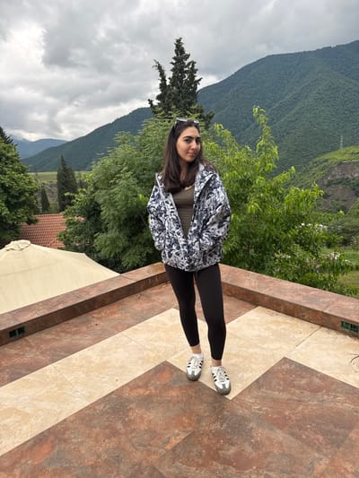 A young woman stands on a tiled patio overlooking a verdant valley and forested mountains under a cloudy sky. She is wearing a black and white patterned jacket, a dark olive top, black leggings, and white athletic shoes. Her hands are in the pockets of her jacket. She has dark hair and is looking to her right. To the left of the woman, a large, light-colored umbrella is visible, as well as the red-tiled roof of a building. The patio is paved with large square tiles in earthy tones of brown, tan, and cream. The background features lush green trees, a distant power line tower, and the imposing, tree-covered slopes of mountains. The overall scene suggests a scenic overlook or a terrace in a rural or mountainous region, potentially near Akhtala, Armenia, given the context.