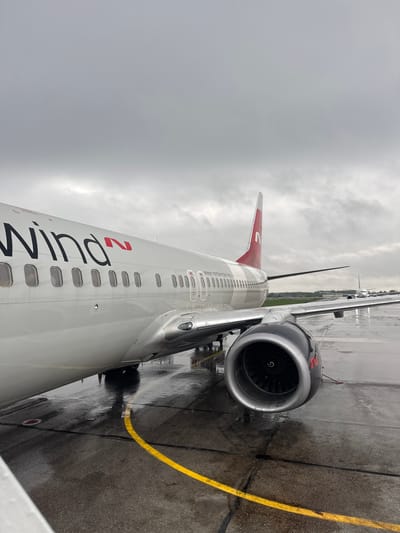 The image shows the exterior of a Nordwind Airlines airplane, possibly a Boeing 737, parked on a wet tarmac. The airplane is white with the airline's name in black and red lettering. Part of the tail fin, also in red and white, is visible. A jet engine is attached to the wing. A yellow line is painted on the tarmac. The sky is overcast, indicating a rainy or cloudy day. The airplane is potentially located at Bolshoye Savino Airport, situated near the city of Bolshoye Savino, Russia. There are no people visible.