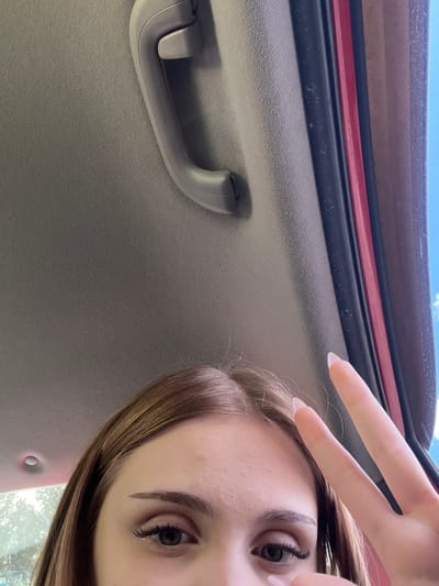 The image shows a close-up, low-angle shot of a young person's face and hand, taken from inside a vehicle, likely in the city of Tolbazy, Russia. The person has light brown hair and is looking towards the camera with one eye visible, the other partially obscured by a hand. Their right hand is raised in a peace sign gesture, with long, manicured fingernails. The interior of the vehicle's roof lining, a grey grab handle, and the edge of a red-painted door frame are visible in the background. The lighting suggests it is daytime, with sunlight filtering in from outside.