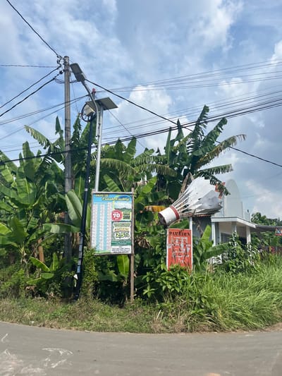 The scene captures a roadside view in Paweden, Indonesia. The main subject is an overgrown area housing signs and infrastructure. A sign advertising land for sale is prominent, bearing the text "DIJUAL Tanah Kavling" and additional information in Indonesian. A smaller sign with the word "PAWEDEN" is also visible. A decorative badminton shuttlecock sits atop one of the structures. The backdrop consists of banana trees and other vegetation. Power lines crisscross overhead, attached to a utility pole equipped with a solar panel and a streetlight. A narrow road borders the overgrown area on the right. A house is visible in the background. The sky is partly cloudy.