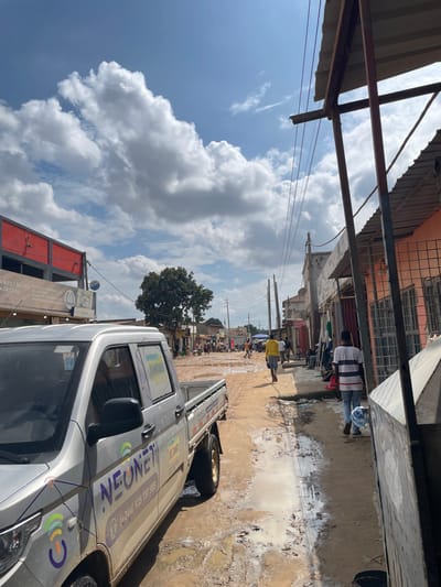 An outdoor scene in Luanda, Angola, captures a bustling unpaved street or alleyway on a partly cloudy day. The ground is muddy and filled with numerous puddles, suggesting recent rainfall. In the foreground on the right, a silver utility truck is prominent, parked or driving along the muddy path. The truck displays "NEONE" in purple lettering, along with the phone number "(+244)935 137 585" and colorful graphic logos. Other visible text on or near the truck includes "MUPS", "MELHOR", "DE CASA", and "GRA".
Overhead, a metal canopy structure with a corrugated roof extends into the frame, with power lines or communication cables visible stretching across the street. In the middle distance, several people are walking along the road; one individual wears a striped shirt, and another a yellow shirt. Buildings of various architectural styles, some featuring red or orange facades, line both sides of the street. A large green tree stands further down the road, and the street appears to continue into the distance with more buildings and activity. The bright sky with puffy white clouds indicates it is daytime.