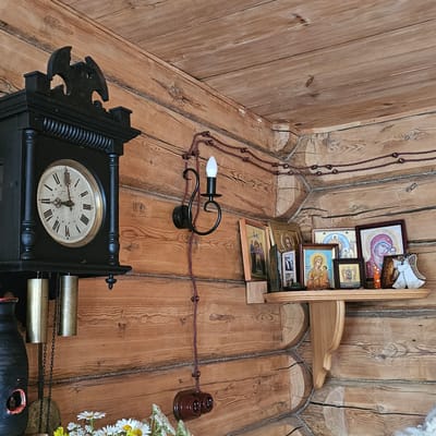 This image depicts an interior scene with rustic wooden walls and flooring, suggesting a dwelling in Vybolovo, Russia. The primary subjects are a collection of religious icons displayed on a wooden shelf, a black grandfather clock, and an antique electric lamp.

The icons, varying in size and framing, are arranged on a light-colored wooden shelf mounted on the wall. They appear to be traditional Orthodox Christian images, with depictions of figures like the Virgin Mary and other saints. Some icons are framed in darker wood, while others are presented more simply. An angelic ornament is attached to the top of the shelf.

A black grandfather clock, with an ornate carved piece on its left side, dominates the lower part of the image. Its face shows Roman numerals and intricate hands, indicating the time is approximately 3:15. Visible on the right side of the clock are two brass weights and chains, suggesting it is a pendulum clock that requires winding.

An old-fashioned electric lamp with a black metal fixture and a white bulb is attached to the wall, connected by a visible, braided wire. This wire runs along the wooden wall and floor, supported by small ceramic insulators. Two similar ceramic electrical outlets are also visible on the wall.

In the lower right corner, a small arrangement of white daisies and other small white flowers adds a touch of natural color to the scene. The lighting suggests it is daytime, with natural light filtering into the room. The overall atmosphere is one of a traditional, perhaps somewhat aged, living space. No visible text is present in the image.