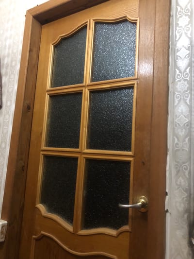 This image shows a close-up, top-down view of a wooden door with glass inserts. The door appears to be part of a home in a settlement in Russia. The glass panes are textured and divided into a 2x3 grid, with decorative wooden trim around each pane. A brass door handle is visible on the right side of the door. Above the door, there is a strip of wallpaper with an ornate, repeating pattern. The overall scene suggests an interior setting, possibly a hallway or entrance. There is no visible text or indication of time of day or weather. The image focuses on the details of the door's construction and design.