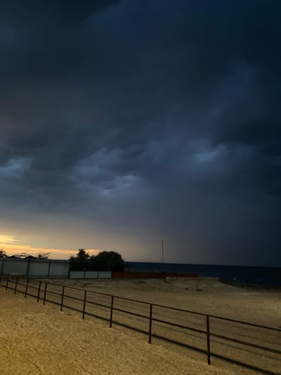 The image depicts a beach scene at dusk in Izberbash, Russia. Dark, ominous storm clouds dominate the sky, casting a heavy shadow over the landscape. To the left, the horizon shows a sliver of yellowish-orange light, suggesting the sun may be setting or has recently set behind the clouds. In the middle ground, there is a sandy beach with a dark metal fence in the foreground. Beyond the fence, there are some low-lying structures and trees. The sea is visible in the distance, appearing dark and choppy under the stormy sky. A few indistinct figures and a blue umbrella can be seen on a boardwalk further down the beach. The overall atmosphere is one of impending weather change.