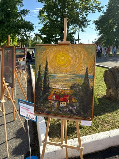 The image depicts an outdoor art exhibition in the city of Progress, Russia. Several paintings are displayed on easels. The central painting, an impressionistic landscape with houses, cypress trees, and a bright yellow sun, is the focal point. In the background, people are seen observing the artworks. The environment includes green trees, a clear sky with some clouds, and a paved walkway. A sign in Russian, identifying the exhibition as "Выставка картин "КРАЙ МОЙ РОДНОЙ"", is visible in the foreground.