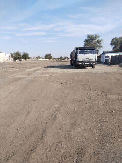 The image depicts an outdoor scene in the city of Ақадыр, Kazakhstan. In the foreground and midground, a dusty, unpaved lot stretches out, marked by tire tracks and scattered debris. A light gray truck, identified by the "KAMA3" logo on its grille, is parked in the midground to the right of the frame. The truck has a dark-colored cargo bed. Behind the truck, several low-rise buildings and trees are visible, suggesting a somewhat rural or semi-developed area. The sky above is blue with scattered white clouds. The overall impression is one of a dry, somewhat desolate environment.
