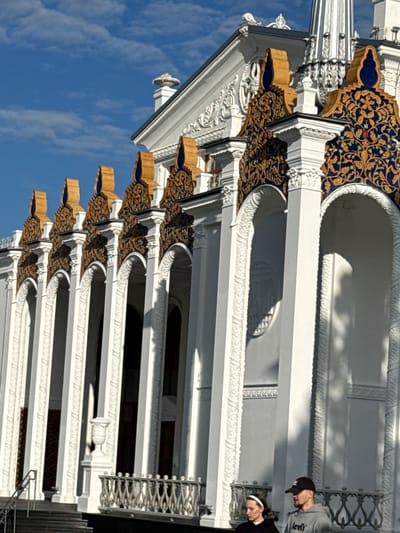The image captures a bright, sunny day with a clear blue sky filled with wispy clouds. The scene focuses on the ornate exterior of a building, likely a pavilion or exhibition hall, characterized by white columns, arched entrances, and elaborate decorative elements. These decorations feature rich blue and gold patterns with intricate floral motifs, topped with golden finials.
In the foreground, two individuals are visible. A woman on the left wears a black hoodie and a white headband. A man on the right wears a grey hoodie with "Levi's" branding and a black baseball cap. They are walking or standing near the building, seemingly interacting with each other. The architecture suggests a classical or neoclassical style, possibly with Eastern influences due to the decorative patterns. The building's façade is highly detailed, with textured surfaces and sculptural elements. Shadows cast by the sun highlight the architectural features and the presence of the people.
This building is located in Moscow, Russia, and is identified as the Armenian Pavilion at VDNKh (Exhibition of Achievements of National Economy). The VDNKh is a vast exhibition center in Moscow, featuring numerous pavilions showcasing the achievements of various Soviet republics and industries. The Armenian Pavilion, specifically, is known for its distinctive architecture that blends classical styles with Armenian motifs.