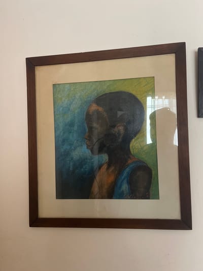 An indoor scene in Bernal Este, Argentina, presents a framed artwork mounted on a light-colored, possibly off-white, wall. The rectangular artwork is enclosed in a dark brown wooden frame, with a light beige or off-white matboard separating the frame from the central image. The artwork itself depicts a side profile portrait of a young person with dark skin, facing towards the left. The background of the painting transitions from blue on the left to green on the right. The subject appears to be wearing a blue garment over their visible right shoulder. The artwork is covered by a reflective glass panel, displaying reflections that obscure parts of the image, including the partial shadow of an individual on the right side. A portion of another, darker framed object is visible on the wall immediately to the right of the primary artwork.