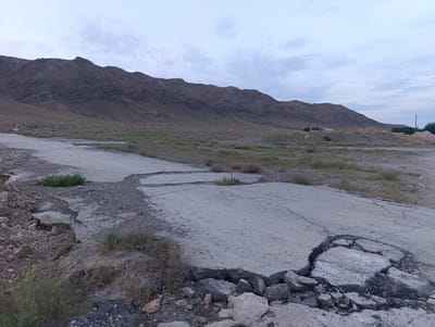 This media file captures a desolate, rural scene in Zarafshan City, Uzbekistan. The primary subject is a severely damaged asphalt road, cracked and broken in multiple places, with large chunks of concrete dislodged and scattered on the surrounding dry, rocky ground. Sparse, dry vegetation grows in patches, struggling to survive in the arid environment.
In the background, a large, barren mountain range dominates the landscape, its slopes etched with the marks of time and erosion. The sky above is overcast, with soft, diffused light suggesting either early morning or late evening. The overall atmosphere is one of neglect and decay, with the crumbling road serving as a stark symbol of a forgotten or abandoned place.
There are no people visible in the image, and no discernible activity is taking place. The notable details include the rugged texture of the mountain, the cracked and broken pavement, the dry, sparse vegetation, and the muted, cloudy sky. The location cues, such as the dry, mountainous terrain and the style of distant, nondescript buildings, are consistent with a rural or semi-arid environment in Central Asia. The overall emotion evoked by the image is one of isolation and perhaps melancholy, due to the signs of disrepair and the vast, empty landscape. No visible text is present in the image.