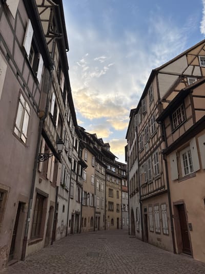 The image depicts a narrow cobblestone street in Colmar, France, lined with traditional Alsatian half-timbered houses. The architecture is characterized by wooden beams forming geometric patterns on the facades, with plaster infill in varying shades of beige, brown, and pink. Many of the windows have white shutters, some with heart-shaped cutouts.

On the left side of the street, the buildings are closer to the viewer and appear to be older, with some signs of wear on the plaster. A wrought-iron street lamp with an ornate design is mounted on one of these buildings. The street itself is paved with uneven cobblestones.

On the right side, the buildings recede into the distance, creating a sense of depth. These buildings also feature the characteristic half-timbered style.

The sky above is partly cloudy with soft, diffused light, suggesting either early morning or late afternoon. The clouds are tinged with hues of yellow and orange, indicating the presence of sunlight filtering through. No people are visible in the scene. The overall atmosphere is quiet and historic, evoking a sense of old-world charm.
