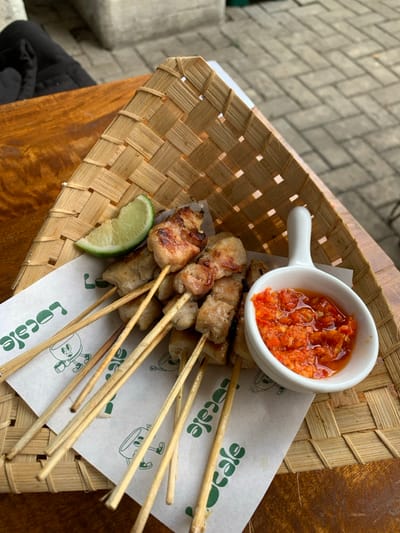 The image displays a meal served outdoors or in a semi-outdoor setting at Locale Cafe, located in Kota Batu, Indonesia. The central subject is a serving of Sate Taichan, consisting of approximately twelve skewers of grilled white-meat chicken, presented on a rectangular woven bamboo basket. The basket is lined with white parchment paper featuring a repetitive green "Locale" logo with a cartoon character graphic. A single lime wedge is positioned on the paper among the skewers. Adjacent to the skewers is a small, white ceramic bowl with a handle, containing a coarse, vibrant red chili sambal (paste). The entire serving is placed on a polished, reddish-brown wooden table. In the background, out of focus, light grey interlocking concrete pavers are visible, indicating an outdoor patio or pathway. A dark fabric object and a light-colored woven structure are partially visible in the upper-left background. No individuals or active interactions are directly depicted.