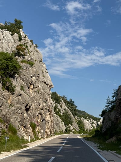 The image depicts a winding asphalt road cutting through a mountainous landscape. On the left, a sheer, grey rock face rises dramatically, dotted with green foliage. On the right, a similar, though less imposing, rock formation is visible, also covered in vegetation. The road is marked with white lines, indicating lanes. The sky above is a clear, vibrant blue, with scattered white clouds. The scene appears to be in a rural, natural environment, likely a mountain pass or a scenic route, and is consistent with the geography of Dragalj, Montenegro, known for its rugged terrain and coastal mountains. No people are present in the image.