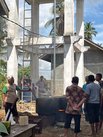 The scene depicts a community event in Rheng, Indonesia, centered around a large cooking operation. A group of men are gathered around a large black vat, which is set over a fire. One man is actively stirring the contents of the vat with a long wooden pole. Another man is seen on the periphery near a wooden table. Four men stand, facing the vat, observing the cooking process.
The cooking operation is taking place beneath the unfinished concrete framework of a building. Palm trees are visible around the structure, and a partly cloudy sky is overhead.