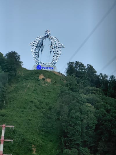 This is a medium shot, slightly angled, of a large, metal, arch-shaped sculpture resembling a stylized person or an abstract figure. The sculpture is illuminated with blue lights and sits atop a grassy, green hillside. The words "РОССЕТИ" are visible in blue letters on a white banner below the sculpture, also illuminated.

The sculpture has a spherical element at the top, resembling a head or a ball, and two large, curved arm-like structures that extend downwards and outwards. The entire structure is made of intricate metalwork, with diagonal beams and cross-bracing visible. It appears to be an outdoor art installation or monument.

The hillside is densely covered with lush green vegetation, predominantly trees and shrubs, indicating a natural, possibly mountainous or hilly, environment. There are some patches of exposed earth or rock on the slope. In the lower left corner of the image, a section of a red and white striped pole, likely part of a power line pylon or a ski lift structure, is visible, with wires extending from it.

The sky is a pale, overcast blue, suggesting a daytime scene with diffused light. The overall impression is of a clear, temperate day. The composition and the subject matter, a prominent monument against a natural backdrop, hint at a tourist attraction or a landmark in a scenic location. The text "РОССЕТИ" identifies the entity associated with the sculpture, which is a Russian energy company. The location context of Vysokoye, Russia, supports the interpretation of a landmark in that region. No people are visible in the frame. The image appears to have been taken from a moving vehicle or a cable car, as suggested by the slight diagonal lines of what might be cables or wires in the upper right and left portions of the image.