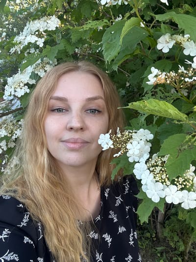 A frontal portrait captures an adult female situated outdoors in Urmanets, Russia. The woman has long, wavy blonde hair, parted slightly off-center, cascading over her shoulders and chest. Her fair skin displays numerous freckles across her nose and cheeks. She possesses light-colored eyes, appearing greenish-grey or hazel, and her expression is neutral with a slight upturn at the corners of her lips. Subtle eye makeup, including a light shimmer, is visible on her eyelids. She wears a black garment, likely a dress or top, patterned with small white botanical sprigs. A thin, rose gold-toned chain necklace is visible on her right clavicle.

The subject is framed by dense green foliage and clusters of white flowers from a flowering shrub, consistent with a *Viburnum* species. These flowers feature larger, flat, four-petaled outer florets surrounding smaller, central fertile flowers. Many of the shrub's green leaves exhibit insect damage, characterized by small holes and irregular edges. A single white flower petal rests on her right cheek. The background consists of additional, slightly blurred green vegetation and white blossoms, suggesting a natural, garden-like setting. The lighting is natural and even, illuminating the scene from the front or slightly above. The woman is looking directly at the viewer.