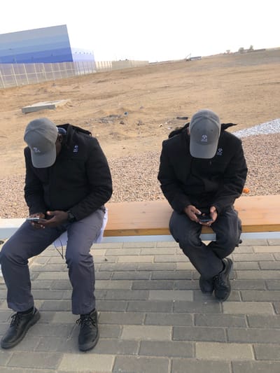 Two men are seated side-by-side on a wooden bench with a light metal frame, positioned on a paved area composed of rectangular grey pavers. Both individuals wear dark-colored padded jackets, dark trousers, and dark lace-up shoes. Each man has a grey baseball cap with a circular logo featuring a triangular/gear-like emblem and text (partially visible, resembling "Saud HVAC" or similar) on the front, and the same logo is present on the left chest of their jackets. Both men are looking downwards, engrossed in their respective smartphones held in their hands. The man on the left has a dark-colored watch on his left wrist and a white earbud wire is visible near his lap.
Behind the bench, the ground transitions to a coarse gravel area, followed by extensive, undulating sandy terrain indicative of a desert environment. In the background, a large building or structure with horizontal blue and light blue panels is visible. A chain-link fence runs in front of this structure. Scattered concrete elements are present on the sandy ground, and further in the distance, what appears to be construction equipment can be faintly discerned. The sky is a light, clear color, suggesting daylight in Hanak, Saudi Arabia.
