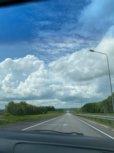 The image shows a view from the dashboard of a car driving on a highway. The road stretches out ahead, with a clear sky filled with dramatic white and grey clouds above. On the right side of the road, there's a metal guardrail and a line of green trees. There are streetlights along the highway. In the distance, a red truck is visible. The foreground shows the car's dashboard. The scene appears to be in a rural area, potentially on the outskirts of Uchkhoza Sel'khoztekhnikuma, Russia, given the highway infrastructure and natural landscape.