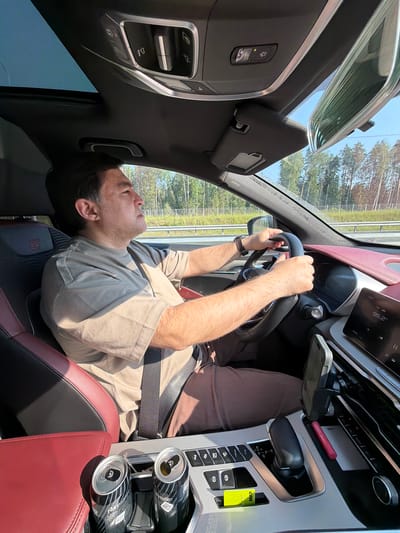 An adult male is driving a car. He is wearing a tan t-shirt and brown pants. He has his hands on the steering wheel. The car has a black interior with red accents. There are two cans of energy drink in the cup holder. The car is driving on a highway with trees visible on the side. The sun is shining.