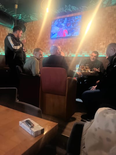 A group of men gather around a table in a bar. Some are watching a sporting event on a nearby TV. A man in a black jacket with white stripes smokes a cigarette. A lighter is on the table. The bar has a textured wall with green and blue neon lighting.