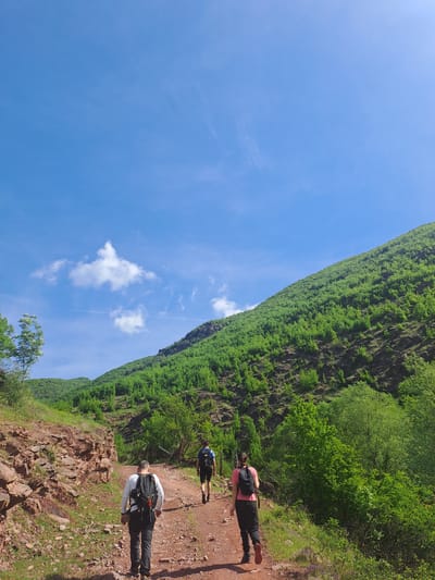 Three people hike on a dirt path beside a cliff face covered in green trees and vegetation. The path is rocky on the left side. The sky is blue with a few clouds. The hikers appear to be in Zverino, Bulgaria. The person in the front is wearing a white shirt, black pants, and a black backpack. The person in the middle is wearing a blue shirt and black pants with a black backpack. The person in the back is wearing a pink shirt and black pants with a black backpack. All are walking.