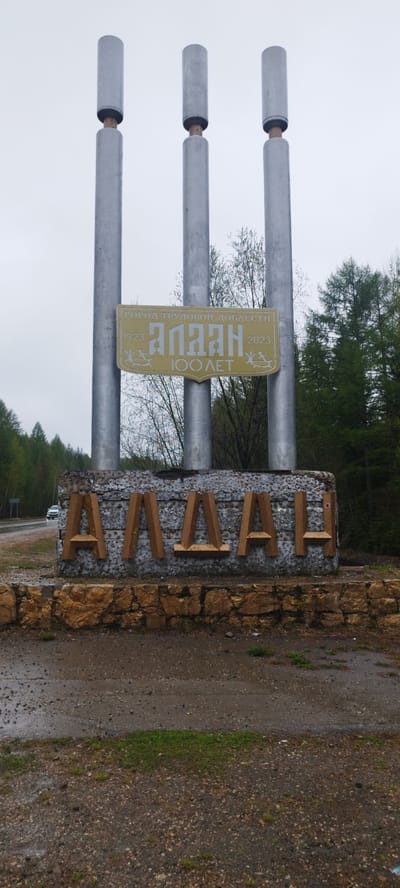 The image shows a sign marking the entrance to the city of Aldan, Russia. The sign consists of three vertical metal columns topped with cylindrical caps. A gold sign is situated between the columns displaying the text "ГОРОД ТРУДОВОЙ ДОБЛЕСТИ 1923 Алдан 2023 100 ЛЕТ," which translates to "City of Labor Valor 1923 Aldan 2023 100 Years." Underneath the gold sign, large wooden block letters spelling "АЛДАН" are embedded in a rough stone structure. The base of the sign is constructed from larger, brown-toned stones. The environment appears to be somewhat overcast and possibly wet, with a road visible to the left and green trees in the background. A white car can be seen driving on the road to the left of the sign. The ground around the sign is a mix of dirt, gravel, and sparse patches of grass.