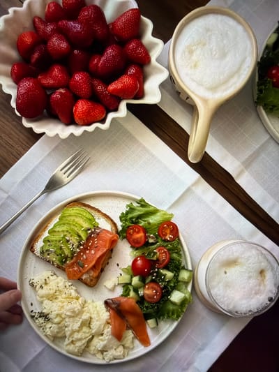 An overhead shot captures a beautifully arranged breakfast or brunch spread on a light-colored, textured tablecloth, set upon a dark wooden table. A central white ceramic plate holds a diverse meal: a slice of toasted bread topped with sliced avocado, a white spread (likely cream cheese), and various seeds (sesame, flax, poppy, or similar). Beside the toast are several slices of smoked salmon and a generous portion of fluffy scrambled eggs, also sprinkled with dark seeds. A small side salad of green lettuce, halved cherry tomatoes, and diced cucumber completes the plate.

To the left, a white ceramic bowl with a decorative wavy rim is brimming with fresh, ripe red strawberries. Two frothy beverages are visible: one in a light-colored ceramic mug or pitcher with a handle, and another in a clear glass, both topped with a thick layer of white foam, suggesting coffee or milk-based drinks like lattes or cappuccinos. A silver metal fork rests on the tablecloth, pointing towards the main plate. A hand, likely adult, is subtly visible in the bottom right corner, suggesting interaction with the table setting. The overall presentation is neat and appealing, indicative of a leisurely meal enjoyed during the daytime. No specific location cues within the image point to Ros Na Rí, Ireland, other than the provided context. There is no visible text in the image.