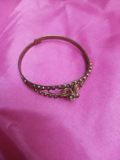 One tarnished gold-tone metal adjustable bangle bracelet is centrally depicted on a vibrant pink fabric surface. The bracelet, approximately 6-8 cm in diameter, exhibits an antique brass or bronze appearance with signs of wear. A distinct hinged mechanism is visible on the upper left side of the bracelet's plain band. Approximately one-third of the bracelet's circumference is adorned with an intricate, openwork filigree design, featuring two prominent intertwined spiral motifs. Along the edges of this decorated section, small, spherical protrusions are uniformly spaced. A single, small, clear, faceted round stone, approximately 2-3mm in diameter, is centrally set within a four-prong claw resembling a floral pattern on the decorated portion. The underlying fabric, possibly satin or silk, displays minor creases and folds, providing a uniform backdrop. The scene is evenly lit, with soft illumination. This scene is located in Qarabucaq qəsəbə, Azerbaijan.