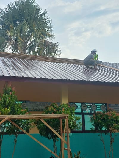Two construction workers are on the roof of a building. The worker on the left is wearing a grey shirt and dark pants, and is crouched down while working. The worker on the right is wearing a yellow safety vest over a grey shirt and a white hard hat. They are working on a corrugated metal roof. A tall palm tree is visible behind the roof, and the sky is cloudy. Below the roof, a portion of a building with yellow walls is visible. In front of the building, there are green bushes with red leaves. There is a wooden structure in the foreground. Text is visible on the building wall below the roof, and some appears to be in Arabic script. The scene appears to be in Buketseuraja, Indonesia, as the text is written in Malay, which is spoken in Indonesia.