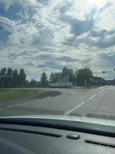 The image shows a road intersection with a large sculpture spelling out "ЧЕЛЯБИНСК" (Chelyabinsk). The sculpture is white and appears to be made of metal or concrete. It is situated in the center of the intersection, surrounded by greenery and trees. The sky is cloudy, with patches of blue showing through. There are road signs and traffic lights visible, indicating this is a public road. The image appears to have been taken from inside a vehicle, looking out through the windshield. The location is identified as Krugloye, Russia, though the name "Chelyabinsk" on the sculpture suggests the scene is on the outskirts of or approaching Chelyabinsk. There are no people visible in the image.