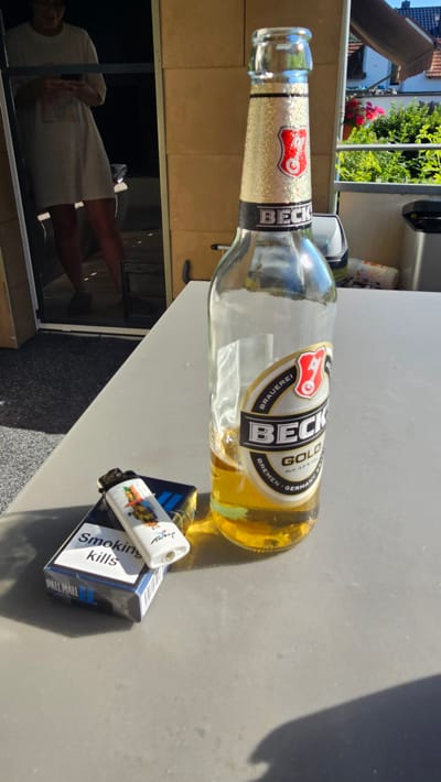 This media file shows a partially empty bottle of Beck's Gold beer lying on its side on a light-colored, textured surface, possibly a table or patio. The bottle has a distinctive glittery gold label around its neck. A pack of Pall Mall cigarettes and a lighter are positioned near the bottle. The lighter has an image of a cartoonish figure on it. The pack of cigarettes displays the warning "Smoking kills" and the brand name "PALL MALL". In the background, a reflection in a glass door or window shows a person wearing a light-colored shirt and shorts, with their legs visible. Outside the window, greenery and what appears to be a building roof are visible, suggesting an outdoor setting during daylight. The overall scene appears to be a casual, possibly domestic setting. The text visible on the beer bottle includes "BECK", "GOLD", "BRAUEREI", "BREMEN GERMANY", and "ALK. 4.9% VOL". The text on the cigarette pack reads "Smoking kills" and "PALL MALL". The time of day appears to be sunny, as indicated by the shadows.