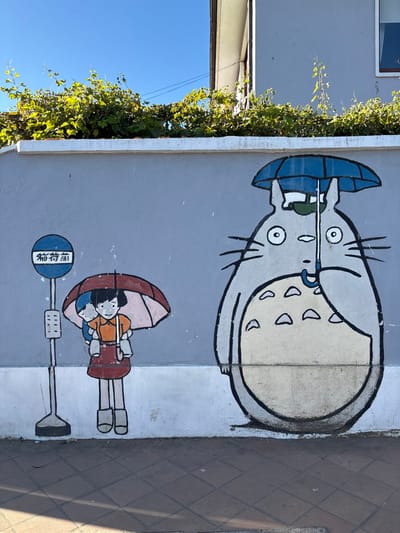 A mural featuring characters from "My Neighbor Totoro" is painted on a light grey wall in Tomé, Chile. On the left, a blue bus stop sign with white Japanese text is depicted. Adjacent to the sign, two animated human figures stand: an older girl in an orange shirt and red skirt carries a smaller child on her back, both sheltered by a partially open red and pink umbrella. To their right, a large grey cat-like creature, Totoro, is illustrated, holding a small blue umbrella while a larger blue umbrella rests over its head. The bottom of the wall has a white stripe and shows some slight discoloration. Above the wall, dense green foliage overhangs, partially obscuring a light grey building with a visible window on the upper right. The scene is illuminated by bright sunlight under a clear blue sky, casting shadows onto the reddish-brown tiled sidewalk in the foreground.