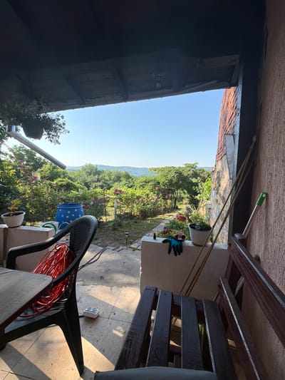 The image depicts a view from a covered patio looking out onto a lush, green landscape with rolling hills in the distance, suggesting a location outside of a dense urban area. The patio itself is furnished with outdoor seating, including a dark plastic chair with a coiled red electrical cord on it and a wooden bench. Gardening tools and potted plants are visible on the patio's edge, and a large blue barrel with white polka dots sits in the yard. The architecture suggests a residential setting. The time of day appears to be morning or late afternoon due to the angle of the sunlight casting shadows. The clear blue sky indicates fair weather. The overall impression is one of a peaceful, rural or semi-rural environment, consistent with the village of Gorno Pavlikene, Bulgaria, known for its natural beauty and agricultural surroundings.