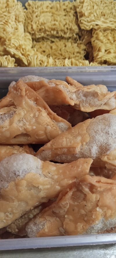 A close-up image shows a clear plastic container filled with a pile of fried, crispy, golden brown samosas. The samosas are layered on top of each other. The container is sitting on a gray surface.  There are stacks of instant noodles, also fried, and crispy in the container above the samosas. The image was taken in Cimaung, Indonesia, at the coordinates -7.074 latitude, 107.547 longitude.  There are no people in the image.