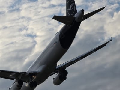 A Lufthansa airplane is captured from a low angle, ascending into a cloudy sky. The plane's tail, adorned with the Lufthansa logo, is prominently featured. The aircraft's registration number, D-AISX, is visible on the fuselage. The underside of the wings and the engines are also visible, with landing gear retracted. The sky is filled with a mix of white and gray clouds. The location is the city of Kelsterbach, Germany, which is situated near Frankfurt Airport, a major hub for Lufthansa.