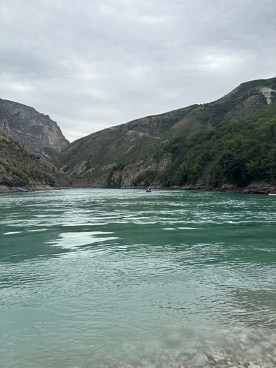 An eye-level, vertical, medium shot captures the expansive turquoise waters of the Chirkey Reservoir in Chirkey, Russia. The reservoir stretches into the distance, framed by steep, rugged mountains on either side. The left mountain is a sheer, rocky cliff face with visible strata. The right mountain is covered in sparse vegetation and trees. The sky above is overcast with grey clouds, casting a diffused light over the scene. Two small boats are visible in the middle distance on the water. The foreground shows the rocky shoreline of the reservoir.