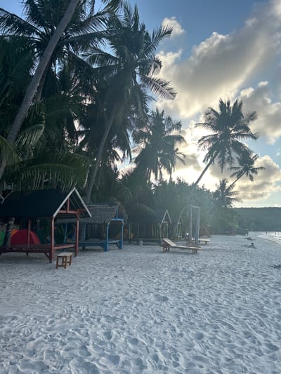 The image depicts a tropical beach scene with white sand, palm trees, and several small huts. The huts are made of wood and thatch, with one having a red and black roof and another a blue base and thatched roof. There are also lounge chairs and swings on the beach. In the background, lush green vegetation lines the shore, leading up to a hillside. The sky is partly cloudy with the sun shining through, creating a bright and atmospheric effect. The scene appears to be a resort or a recreational area. The location is a beach in A R A, Indonesia.