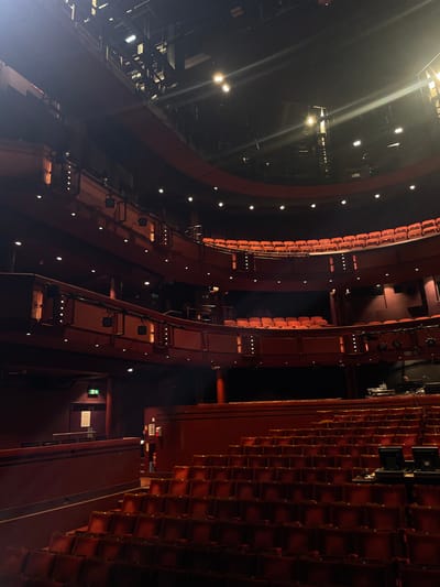 The image shows the interior of the Towngate Theatre in Basildon, United Kingdom. The theatre is empty, with rows of red seats arranged in a semi-circular configuration. The stage area is visible in the distance, with some equipment and lighting rigs present. The lighting is dim, and the atmosphere is quiet and subdued. The walls are a deep red, creating a warm and inviting ambiance. The seats are arranged in a series of balconies. The balconies are empty. There is a door with an exit sign visible on the left-hand side of the image. The door leads to a hallway. There are a few ceiling lights and a few spotlights illuminating the seats and the stage. There are no people visible in the image.