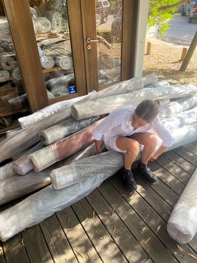 A woman in a white shirt and shorts sits atop a stack of wrapped rugs or carpets on a wooden deck. She's wearing dark shoes and appears to be inspecting the rolls. Adjacent is a wooden-framed glass display showing more rugs rolled up inside. The scene is outdoors, likely near a rug store or warehouse, under bright daylight.