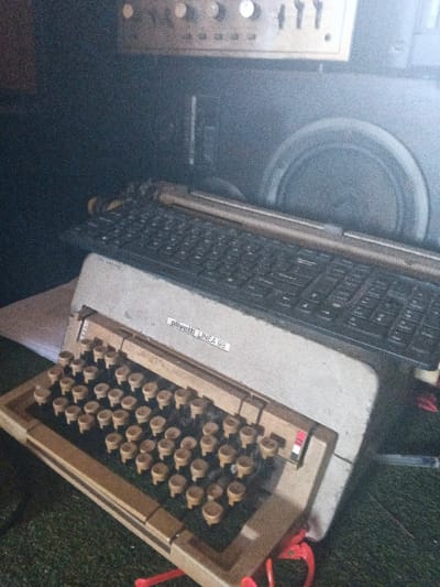 The image depicts a static indoor scene in Dogsenang Gorphoog, Bhutan, featuring multiple electronic and mechanical devices. In the foreground, an Olivetti Linea 98 mechanical typewriter is positioned, characterized by its beige, rounded keycaps and a prominent label "olivetti LINEA 98" on its beige chassis. A red ribbon or cord is visible at the lower right base of the typewriter. This typewriter rests on a dark green, textured surface, possibly carpet or artificial turf. Immediately behind the Olivetti, a modern black computer keyboard with flat, rectangular keys is partially visible. The background is dark, revealing audio equipment. An electronic receiver or amplifier with multiple silver knobs and switches is visible at the top. Below this, a large circular speaker cone is partially visible within a dark enclosure. The lighting is dim, casting shadows and contributing to a grainy image quality. No individuals are present, and no active interactions are depicted.