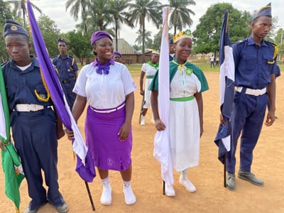 In a seemingly rural area of Tabu, Nigeria, a group of people, mostly young, are standing on a dirt-covered field. The individuals are clad in uniforms, suggesting they belong to an organization or group.
Two young men on the left and right of the frame are in matching blue uniforms with white belts and caps, holding flags with green, white, and purple colors.
Two young women stand in the foreground, one wearing a white blouse, purple skirt, and matching beret, and the other in a white dress with green accents and a crown. Both carry flags, one predominantly white and the other purple.
Several other uniformed individuals are visible in the background. The background also features a variety of trees, including palm trees, and rudimentary buildings. The sky overhead is overcast, providing diffused lighting.