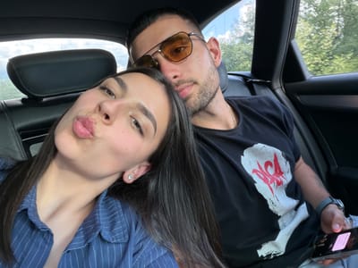 Two individuals are in the interior of a vehicle. One individual wears sunglasses and a gold chain. The other individual is wearing a blue pinstripe shirt. A cell phone is visible in the image. Outside, trees and sky can be seen. The location is unspecified.