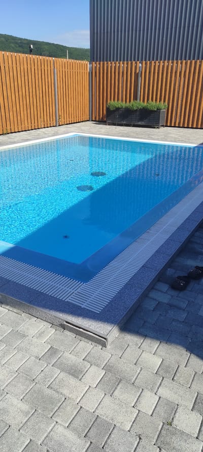 This image depicts a clean, modern outdoor swimming pool area on a sunny day, likely in or near Tulskiy, Russia. The pool itself is a bright, clear blue, reflecting the sky. It is surrounded by a paved patio made of grey interlocking bricks. A low, slatted drainage system runs along the edge of the pool. To the left and behind the pool, a light brown wooden fence with vertical slats encloses the area. Beyond the fence, a lush green hillside is visible under a bright blue sky. To the right of the pool, a dark grey, corrugated metal building stands. In the background, a rectangular black planter filled with green plants is placed against the fence, near the building. A pair of dark flip-flops rests on the paved area near the bottom right corner of the image. The scene is tidy and appears to be a well-maintained private residence or resort.