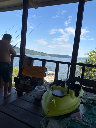 The image depicts a serene outdoor scene on a wooden porch overlooking a calm body of water. The location is identified as Zvanika, Bulgaria. In the foreground, a person, shirtless and wearing dark shorts, stands near a wooden railing, facing away from the camera and towards the water. Several fishing rods are positioned in front of the person and are angled towards the sky, suggesting an ongoing fishing activity.
On a wooden table in the mid-ground, a bright orange toolbox is open, revealing various tools and supplies within. Next to it sits a large, translucent plastic container. A lime-green bait boat, equipped with what appears to be an antenna, is positioned prominently on the table, also facing the water. A patterned cloth is draped over the edge of the table beneath the bait boat.
The background reveals a wide expanse of water, with a distant shoreline characterized by rolling hills and a scattering of buildings. A small boat with a tan canopy is visible on the water. The sky above is a vibrant blue, dotted with fluffy white clouds. The overall atmosphere is one of relaxation and outdoor recreation, likely associated with a fishing trip.