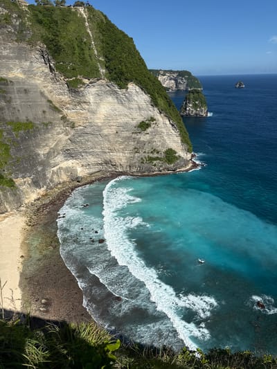 The image depicts an aerial view of a coastal landscape in Bunga Mekar, Indonesia. A large, vertical cliff face dominates the left side of the frame, covered in vegetation at the top and revealing geological layering towards the bottom. To the right of the cliff, a beach stretches along the shoreline. Waves crash against the shore, creating a white foam pattern on the turquoise water. The ocean transitions from a lighter turquoise near the shore to a darker blue further out. In the distance, a few small, rocky islands rise from the sea. The sky is a clear, light blue.
