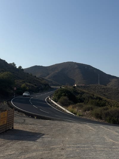 The image shows a winding asphalt road cutting through a hilly, arid landscape under a clear blue sky. A white sedan is traveling on the road, following its curves. The roadside is lined with sparse vegetation, including bushes and small trees. A metal guardrail runs along the outer edge of the road. In the background, a large, undulating hill dominates the scene, with a few scattered buildings and a cell tower visible on its slopes. In the foreground, a wooden barrier with yellow signage is present, indicating "PROTECCIÓN CIVIL PLAYA NO VIGILADA 112" which translates to "Civil Protection Unsupervised Beach 112". This suggests the location is near a coastal area in La Unión, Spain, specifically indicating emergency services information for an unsupervised beach. The overall impression is one of a rural, scenic route.