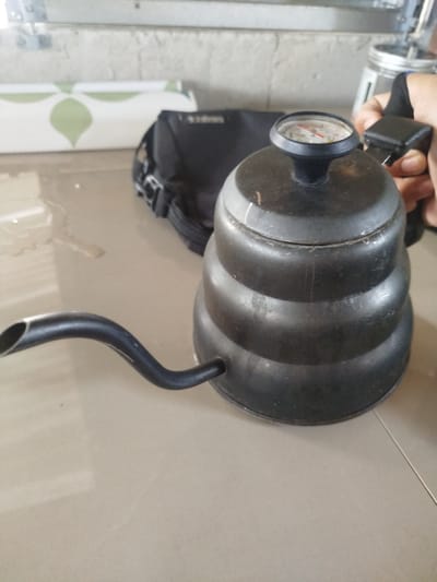 A black kettle with a built-in thermometer sits on a light brown tiled surface. The kettle has a narrow spout and a tiered, bulbous body. The thermometer on top shows a reading in red. A person's hand is visible holding the kettle's handle. In the background, a black bag and a roll of patterned material are present. The scene appears to be indoors, on a tiled floor, likely in Dukupuntang, Indonesia.