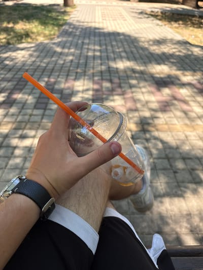 A close-up, POV shot shows a person's hand holding a clear plastic cup with an orange straw and ice cubes. The person is wearing a watch with a black leather band and a silver buckle. They are also wearing black shorts with white trim and white socks with athletic shoes. The background shows a paved walkway with shadows cast by trees. The image is taken outdoors during the day. The location is specified as the city of Gresovsky, Ukraine.
